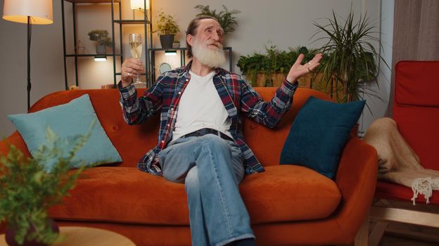 Joyful senior old grandfather hold glass of champagne cheering, drinking, celebrating success win, career grow up, lucky situation at home in room. Mature man celebrating rent or purchase of new house