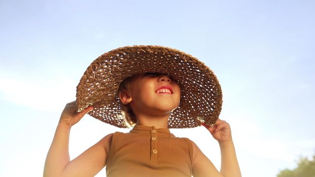 Adorable smiling boy in straw hat on blue sky background. Funny child with kind face looking to camera, enjoying summer outdoors. Childhood, countryside leisure. High quality photo