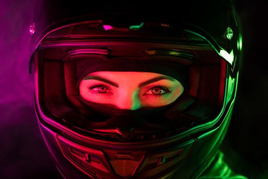 Portrait of confident motorcyclist woman in closed motorcycle helmet. Young driver biker looking to camera under pink and green neon colorful light at night. High quality photo