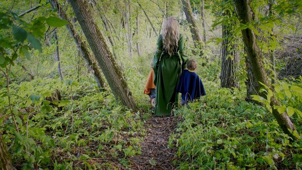 Two little toddler boys cosplay gnomes or hobbits with elf woman walking in green forest. Unrecognizable fairy tale characters. Halloween, kids concept. Amazing light. High quality photo