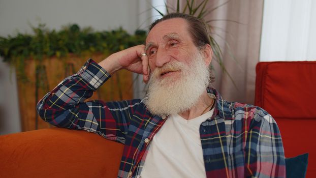 Close-up of happy calm senior old grandfather man pensioner smiling friendly, glad expression looking away dreaming resting, relaxation feel satisfied concept good news sitting on sofa at home room