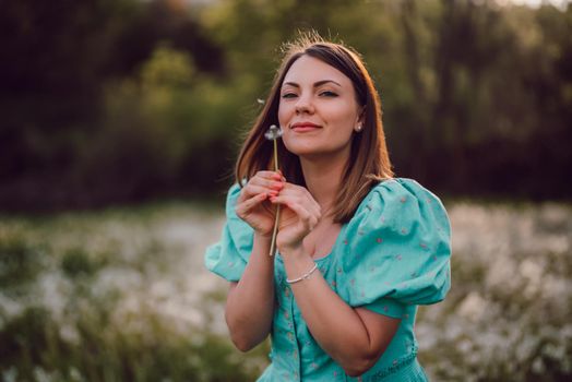 Smiling woman beautiful blowing on ripened dandelion in park. Girl in retro turquoise dress enjoying summer in countryside. Wishing, joy concept. Springtime, aesthetic portrait. High quality photo