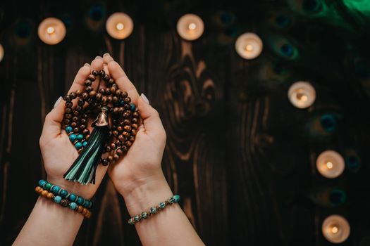 Woman holds in hand wooden mala beads strands used for keeping count during mantra meditations. Weaving and creation. Wooden background with candles and feathers. Spirituality, religion, God concept