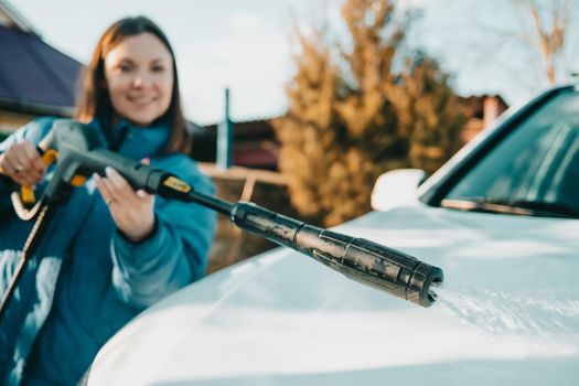 Manual car wash with jet of high pressure water. Woman washes white automobile outside on territory of private house. Caring for vehicle concept. High quality photo