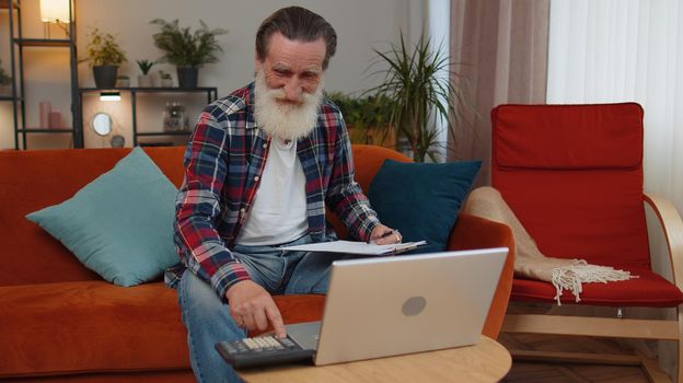 Happy smiling senior grandfather man accounting payments and calculating monthly expenses bank bills, analysing paperwork at home room couch. Elderly freelancer working online remote job on laptop
