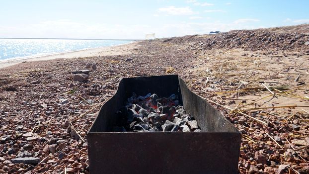 A large barbecue on the shore of the lake, there is smoke from the coals. Bright sunny day, blue lake water, yellow sand with dry branches. Preparation of coals for meat. Food in nature. Lake Balkhash