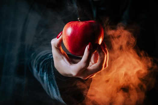 Woman as witch in black offers red apple as symbol of temptation, poison. Fairy tale, white snow wizard concept. Spooky halloween, cosplay. Smoke, haze background. High quality photo