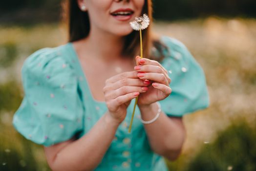 Smiling woman beautiful blowing on ripened dandelion in park. Girl in retro turquoise dress enjoying summer in countryside. Wishing, joy concept. Springtime, aesthetic portrait. High quality photo