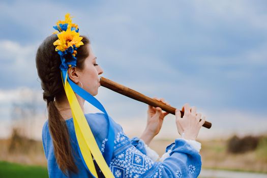 Woman playing woodwind wooden flute - ukrainian sopilka outdoors. Folk music, culture concept. Musical instrument. Lady in traditional embroidered shirt - blue vyshyvanka. High quality photo