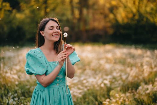 Happy woman beautiful blowing on dandelion in park. Girl in vintage blue dress. Wishing, joy concept. Springtime, aesthetic portrait. High quality photo