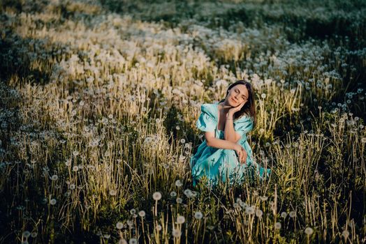 Attractive woman sitting on ripened dandelion lawn in park. Girl in retro turquoise dress enjoying summer in countryside. Wishing, joy concept. Springtime, aesthetic portrait. High quality photo