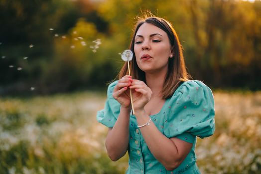 Happy woman beautiful blowing on dandelion in park. Girl in vintage blue dress. Wishing, joy concept. Springtime, aesthetic portrait. High quality photo