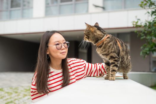 A striped cat sits on a bench outdoors. Young woman walking with her pet outdoors