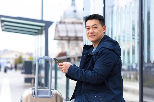 portrait of Asian tourist looking at camera and smiling, happy man arriving in new city, holding phone for online hotel booking and car rental