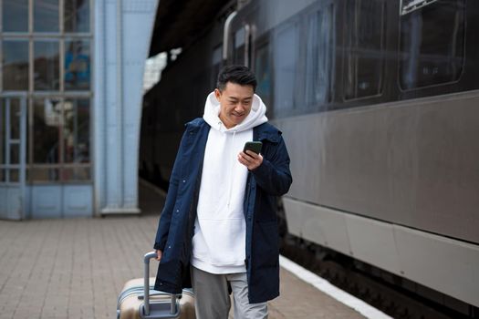 Successful asian man tourist near big train, reading news uses mobile phone, smiling and happy man with big suitcase at train station