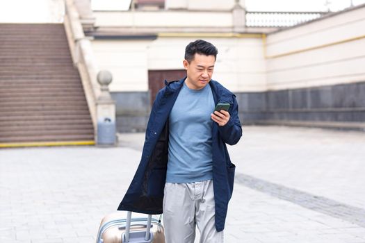 Asian tourist, male passenger arrives from the airport, walks around the city with a suitcase, a man smiles and rejoices uses the phone and the application to book accommodation and book a taxi