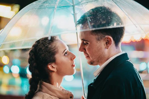 Young beautiful couple spending time together on date in amusement park at night. Rainy weather, autumn. Lovers standing under umbrella. Illuminated background. High quality photo