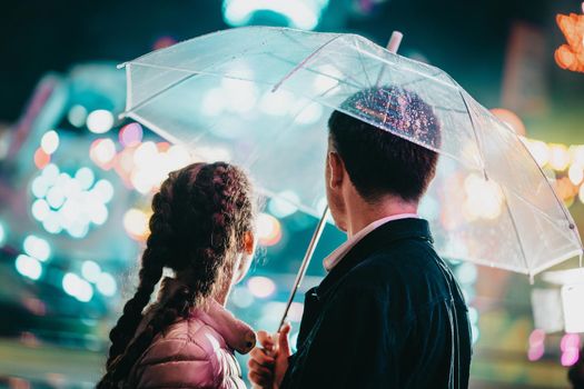 Young beautiful couple spending time together on date in amusement park at night. Rainy weather, autumn. Lovers standing under umbrella. Illuminated background. High quality photo