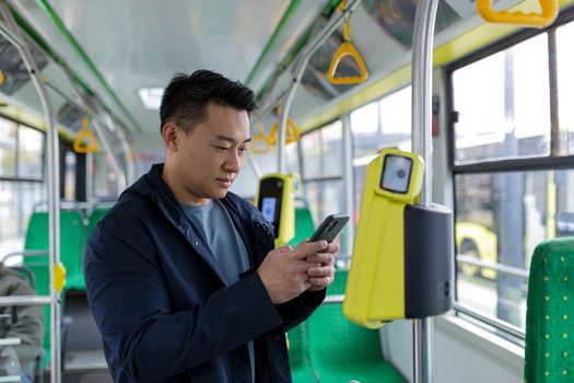 Young asian man corresponds and reads the news online, sitting in the bus, a passenger in casual clothes smiles