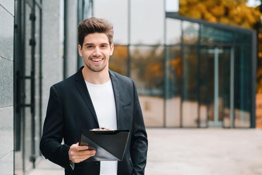 Portrait of young handsome man in jacket with documents, utility bills, report. Businessman near office building. He is satisfied with work of corporate staff. High quality photo