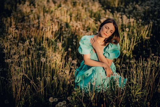 Attractive woman sitting on ripened dandelion lawn in park. Girl in retro turquoise dress enjoying summer in countryside. Wishing, joy concept. Springtime, aesthetic portrait. High quality photo