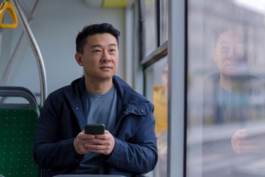 Young asian man corresponds and reads the news online, sitting in the bus, a passenger in casual clothes smiles