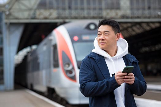 Successful asian man tourist near big train, reading news uses mobile phone, smiling and happy man with big suitcase at train station