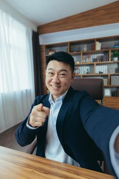 Cheerful and funny Asian businessman looks at the camera of a smartphone, talks on a video call, a man works in the office, waves a congratulatory gesture