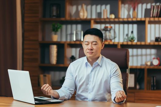 Successful businessman meditates sitting at a table in a classic office, Asian man in shirt relaxes, after working on laptop, man at work