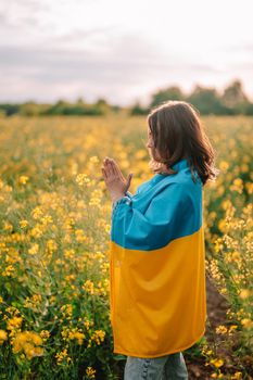 Ukrainian patriot woman praying with national flag in canola yellow field. Pray, stand with Ukraine. Peace, independence, freedom, victory in war. High quality photo