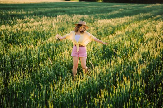 Happy woman in straw hat dancing in fresh green wheat field. Grass background. Amazing nature, farmland, growing cereal plants. Stylish lady with eyewear. High quality photo