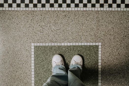 Top view of woman legs in jeans and white sneakers on pattern tile. Copy space, selfie. Black and white ornament, european entrance in public building. High quality photo