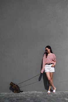 A young woman walks with a gray tabby cat on a leash against a gray wall