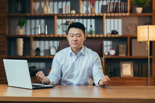 Successful businessman meditates sitting at a table in a classic office, Asian man in shirt relaxes, after working on laptop, man at work