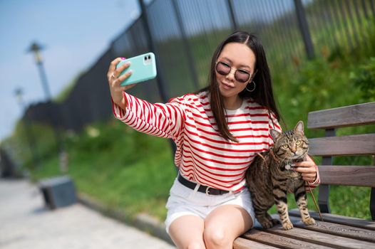 Young woman sits on a bench with a tabby cat and takes a selfie on a smartphone outdoors