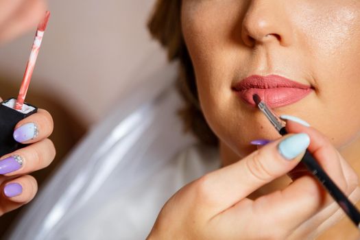 Makeup artist applies red lipstick. Beautiful female face. Hand of a make-up master painting lips of a young girl beauty model. Makeup in progress