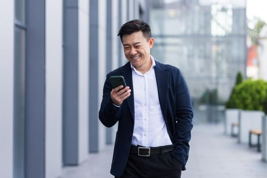 asian male freelancer walking near business center holding phone, smiling reading news, successful businessman