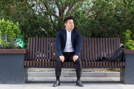 Asian businessman performing breathing exercises trying to calm stress, sitting on a bench during a lunch break in a business suit