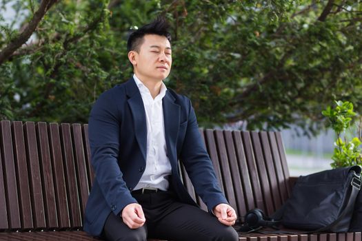 Asian businessman performing breathing exercises trying to calm stress, sitting on a bench during a lunch break in a business suit