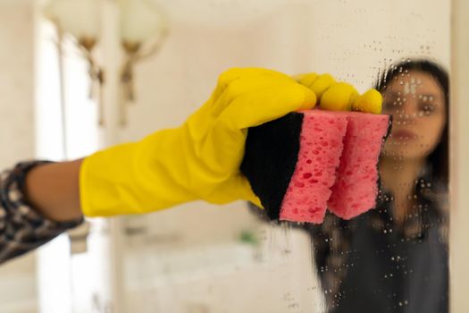 A young girl is cleaning the bathroom, applying detergent with a spray and washing the mirror with a sponge in yellow gloves on her hands. Woman taking care of the cleanliness of her home.