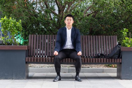 Asian businessman performing breathing exercises trying to calm stress, sitting on a bench during a lunch break in a business suit