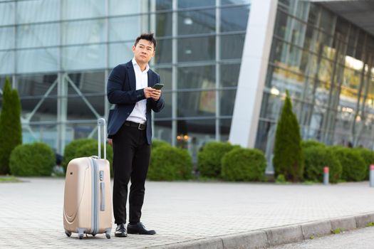 Businessman trying to call a taxi using an app and a mobile phone, asian man at the train station with a big suitcase