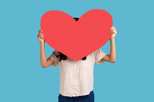 Anonymous date. Portrait of woman hiding her face behind big read paper heart, holding symbol of love, care, generosity, give hope, wearing white shirt. Indoor studio shot isolated on blue background.