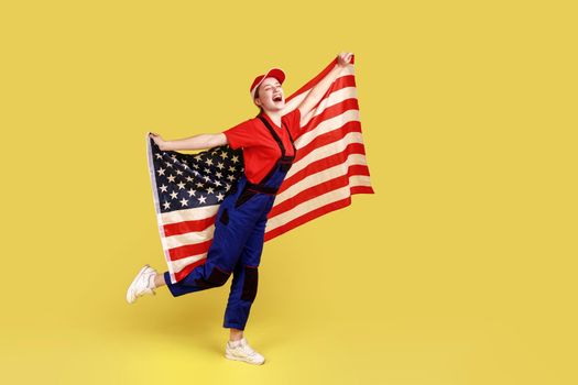 Full length portrait of excited worker woman holding with american flag in hands, pretending she is flying, wearing overalls and red cap. Indoor studio shot isolated on yellow background.