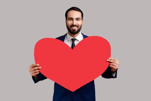 Portrait of smiling handsome bearded man holding big red heart, expressing positive romantic emotions, wearing official style suit. Indoor studio shot isolated on gray background.