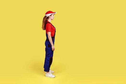 Profile portrait of worker woman standing and looking ahead with positive facial expression, being in good mood, wearing overalls and red cap. Indoor studio shot isolated on yellow background.