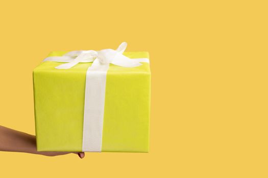 Closeup side view of woman hand holding out yellow gift box with ribbon, giving present on holiday, bonuses and surprises concept. Indoor studio shot isolated on yellow background.