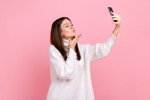 Side view portrait of romantic beautiful female sending air kisses to followers while streaming, wearing white casual style sweater. Indoor studio shot isolated on pink background.