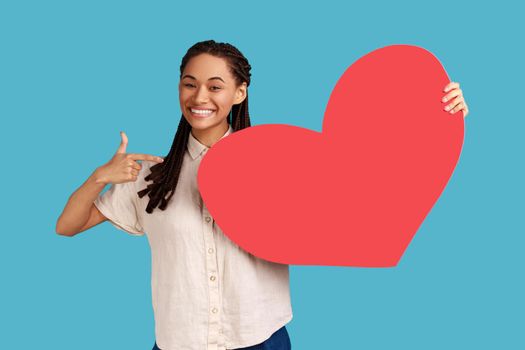 Portrait of enthusiastic woman with black dreadlocks pointing at big red paper heart and smiling, expressing extreme joy, greeting on valentines day. Indoor studio shot isolated on blue background.