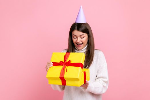 Positive happy girl in party cone looking inside wrapped present box, open gift, pleasant surprise, wearing white casual style sweater. Indoor studio shot isolated on pink background.